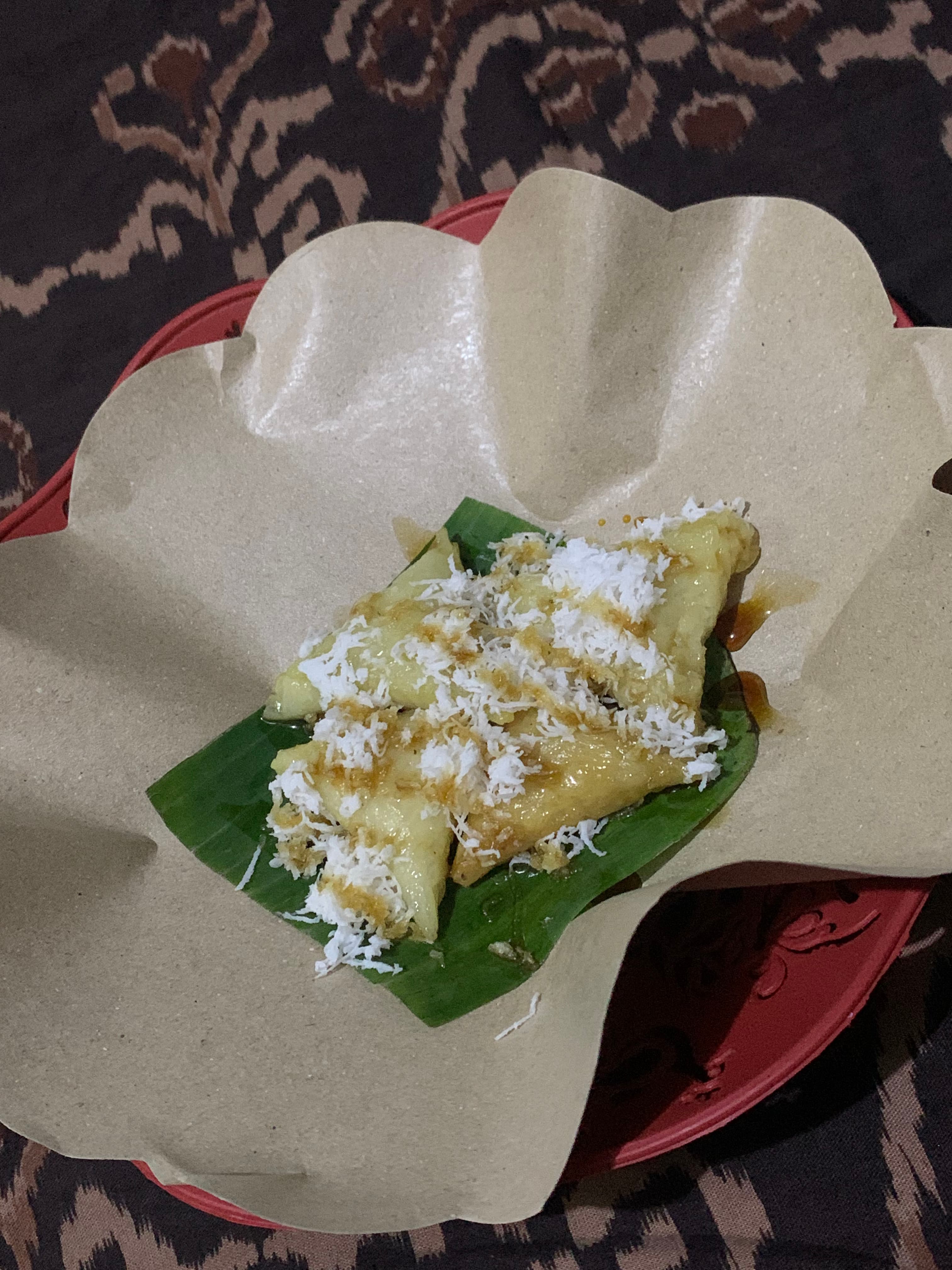 Kue Lukis berbentuk lipatan segitiga dengan taburan kelapa parut dan gula merah cair di atas daun pisang